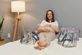 Charming young adult brown haired pregnant woman lying on bed touching her belly looking at camera with toothy smile resting after Royalty Free Stock Photo