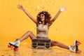 Charming young african woman in retro wear and roller scates throwing confetti while sitting with boombox