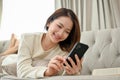 Charming young Asian female laying on her comfy sofa, chatting with her friends on smartphone