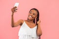 Charming young black woman taking selfie with smartphone, filming content for her video blog on pink studio background