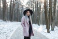 Charming young blond woman in a stylish lilac coat in a black dress in an elegant black hat is standing in a snowy forest