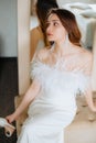 Young bride sitting on sofa in shoulderless wedding dress with feathers, hold shoe in hand. Lady wear tassel earrings.