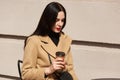 Charming young brunette woman with long dark hair wearing coat, sitting at table in street cafe and drinking coffee or tea, Royalty Free Stock Photo