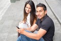 Charming young couple sitting on building steps using mobile phone and interacting happily, urban tourist concept