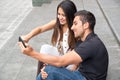 Charming young couple sitting on building steps using mobile phone and interacting happily, urban tourist concept