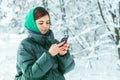 A charming young girl is chatting on social networks and typing messages on her cell phone in the woods on a cold winter day. In t Royalty Free Stock Photo