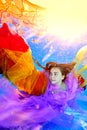 A charming young girl poses underwater in a pool in a purple dress and with a red and yellow cloth in her hands. A