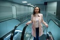 Attractive young woman, passenger in beige coat holds suitcase and smiles cutely looking at camera while going up escalator in the Royalty Free Stock Photo