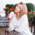 Charming young woman with blue eyes in a white sweater in jeans sits and drinks a sweet drink at a wooden table in a cafe Royalty Free Stock Photo