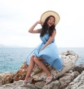 Charming young woman with dark hair sits on a stone next to the sea. Blue dress and straw hat. Fashionable beauty. Royalty Free Stock Photo