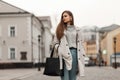 Charming young woman in a fashionable knitted sweater in a chic trench coat in blue jeans with a black bag is standing on a street