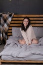 charming young woman in knitted beige sweater in bed with grey linens. modern loft bedroom in brown and gray colors