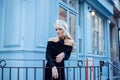 Charming young woman in black silk dress standing on street in Paris. Blondie girl stoped near blue building and smile.