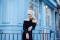 Charming young woman in black silk dress standing on street in Paris. Blondie girl stoped near blue building and smile. Royalty Free Stock Photo