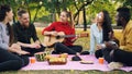 Charming young woman is playing the guitar sitting on blanket with friends on picnic, girls and guys are clapping hands Royalty Free Stock Photo