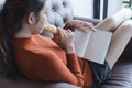 Charming young woman sitting on sofa eating a croissant and reading a book. blank book for graphic montage.