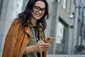 Chatting with friend. Young happy fashionable woman wearing eyeglasses walking on the city street, using her smartphone Royalty Free Stock Photo