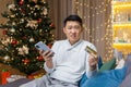 Cheated man at home sitting on sofa and looking at camera, Asian Christmas day frustrated trying to make a purchase in Royalty Free Stock Photo