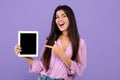 Check this.Cheerful armenian woman holding and pointing at digital tablet with black blank screen, purple background