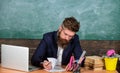 Check homework. Teacher bearded hipster with eyeglasses sit in classroom chalkboard background. Teacher sit desk with