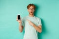 Check this out. Handsome redhead guy in glasses pointing finger at blank smartphone screen, showing online promotion