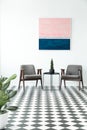 Checkered floor with a plant in a lobby interior with two armchairs, table and painting in the background
