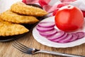 Napkin, fried cheburek in brown dish, plate with tomato and slices of red onion, fork on wooden table