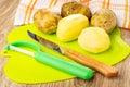 Napkin, unpeeled potatoes, peeler, knife, peeled potato on cutting board on wooden table