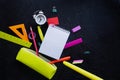 Checkered notepad, alarm clock and multicolored stationery falling out of a bright yellow pencil case against a black background