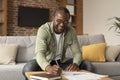 Cheerful adult black businessman in glasses makes notes, studies and works in living room interior, copy space