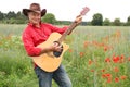 Cheerful adult man in a red shirt and a coboy hat on a field of green poppies plays guitar, posing and having fun, concept musical Royalty Free Stock Photo