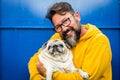 Cheerful adult man smile and hug with love his own old dog pug in a portrait with yellow and blue color - people with animals and Royalty Free Stock Photo