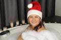 Cheerful adult woman in santa claus hat sits in a big round bath in white foam flakes, imitation of winter snow, christmas concept