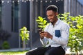 Cheerful african american guy using mobile phone sitting on bench in city park outdoors. Happy black man in casual clothes with a Royalty Free Stock Photo