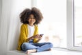 Cheerful african american little girl sitting on windowsill with smartphone Royalty Free Stock Photo