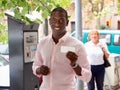 Cheerful African American man showing ticket for car parking purchased in parking meter on city street Royalty Free Stock Photo