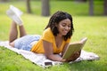 Cheerful African American woman reading interesting book while lying on green lawn at park Royalty Free Stock Photo