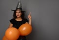 Cheerful African American woman in wizard hat, dressed in black, holds orange inflated air balloons and points on a copy space on Royalty Free Stock Photo