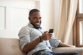 Cheerful african guy sitting on couch spending time using smartphone Royalty Free Stock Photo