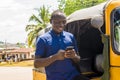 cheerful african man standing next to his tuk tuk taxi smiling and using his smart phone Royalty Free Stock Photo