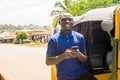 cheerful african man standing next to his tuk tuk taxi smiling and using his smart phone Royalty Free Stock Photo