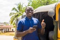 cheerful african man standing next to his tuk tuk taxi smiling and using his smart phone giving a thumbs up Royalty Free Stock Photo