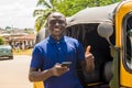 cheerful african man standing next to his tuk tuk taxi smiling and using his smart phone giving a thumbs up Royalty Free Stock Photo