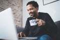 Cheerful African man using computer and smiling while sitting on the sofa.Black guy holding ceramic cup in hand.Concept Royalty Free Stock Photo