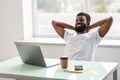 Cheerful african man is watching at his laptop screen, at his work place, with arms behind the head, resting, smiling, in the Royalty Free Stock Photo