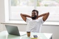 Cheerful african man is watching at his laptop screen, at his work place, with arms behind the head, resting, smiling, in the Royalty Free Stock Photo