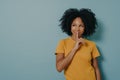 Cheerful afro girl showing shhh sign with finger near lips, standing over pastel blue background