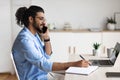 Cheerful Arab Freelancer Guy Talking On Cellphone And Writing Notes To Notepad Royalty Free Stock Photo