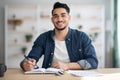 Cheerful arab guy in casual sitting at workdesk, taking notes