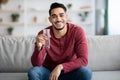 Cheerful arab guy sitting on couch, drinking water, copy space